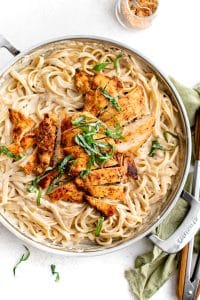 Sliced blackened chicken breasts arranged over creamy fettuccine Alfredo in a stainless steel skillet, topped with fresh basil.