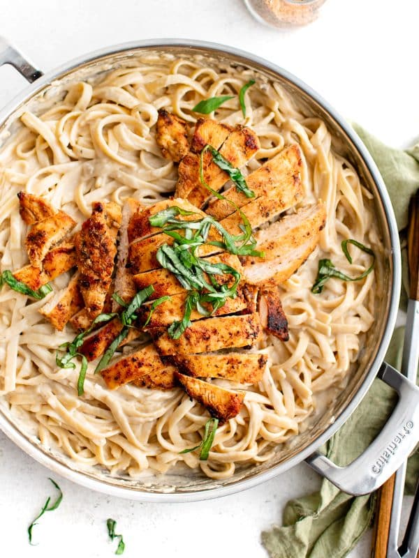 Sliced blackened chicken breasts arranged over creamy fettuccine Alfredo in a stainless steel skillet, topped with fresh basil.
