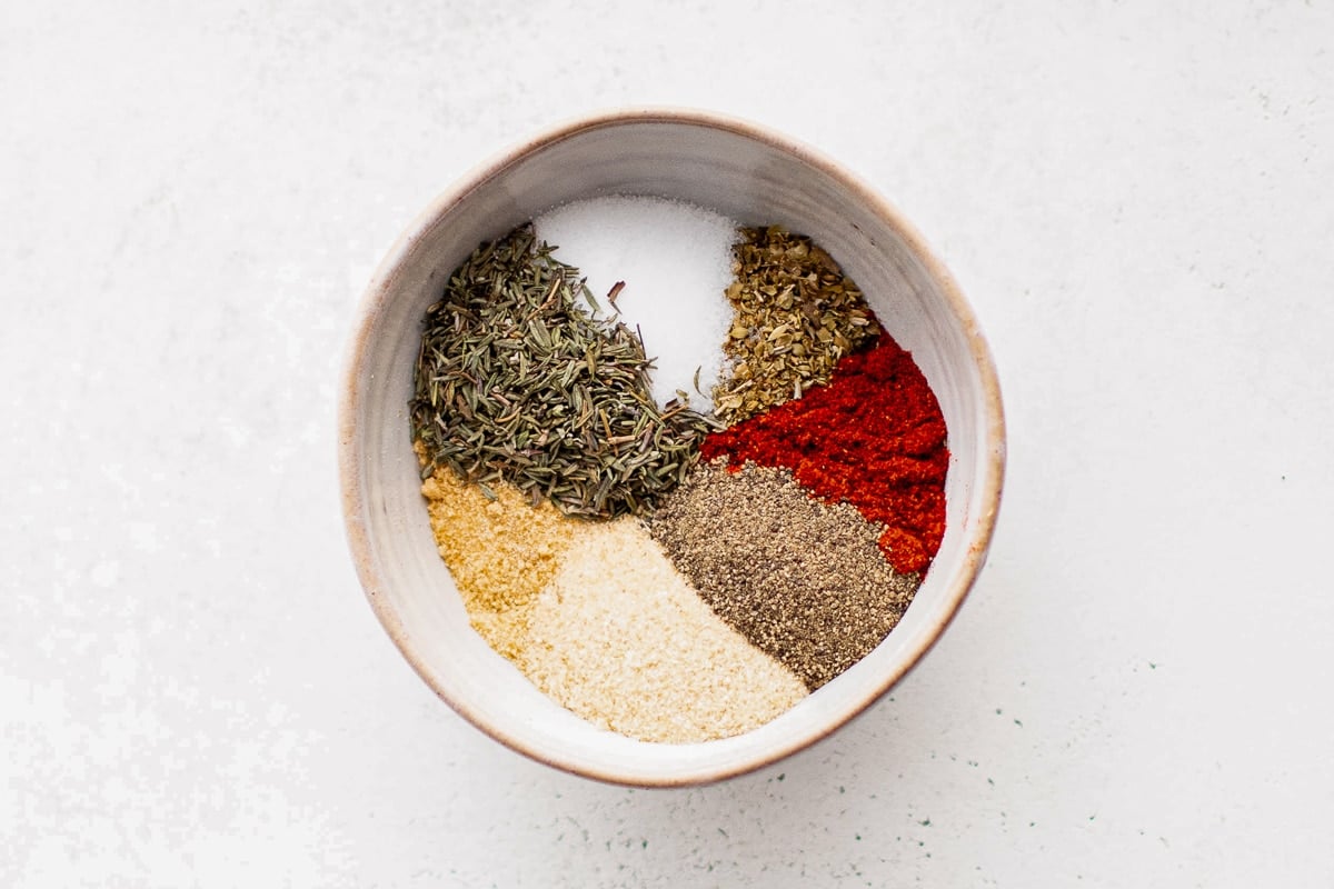 A ceramic bowl containing separated piles of dried spices used to make blackened seasoning. Ingredients include smoked paprika, garlic powder, onion powder, black pepper, salt, thyme, and oregano, artfully arranged on a white background.