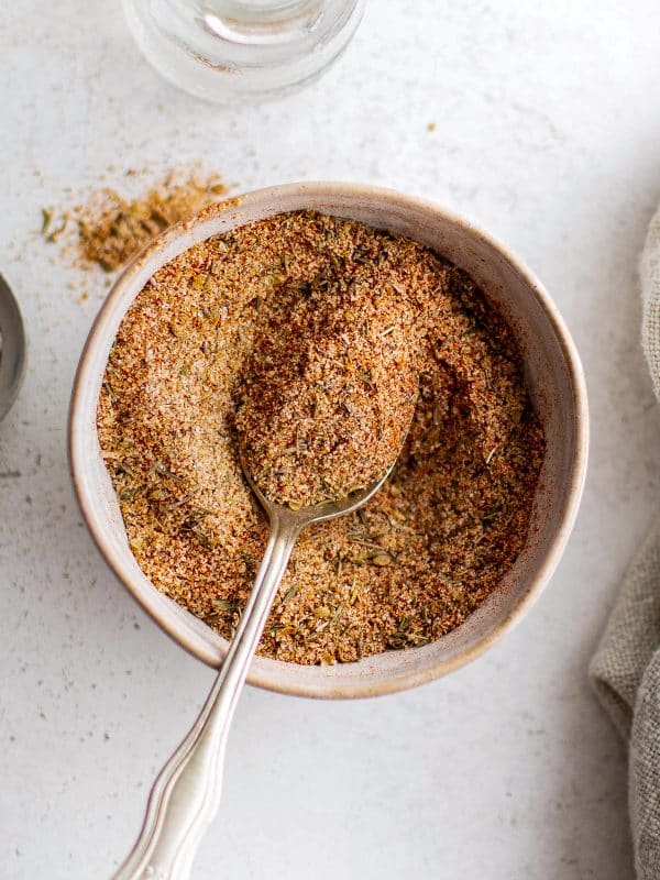 A bowl of freshly mixed blackened seasoning with a spoon resting inside. The spice blend shows a coarse texture with visible herbs.
