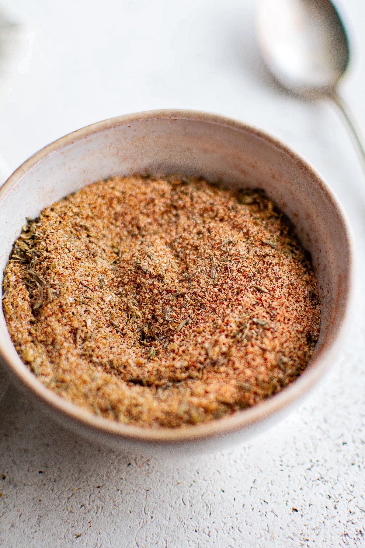 A bowl of fully mixed blackened seasoning displayed on a textured white surface. The blend has a rich reddish-brown hue with visible flecks of dried herbs like thyme and oregano.