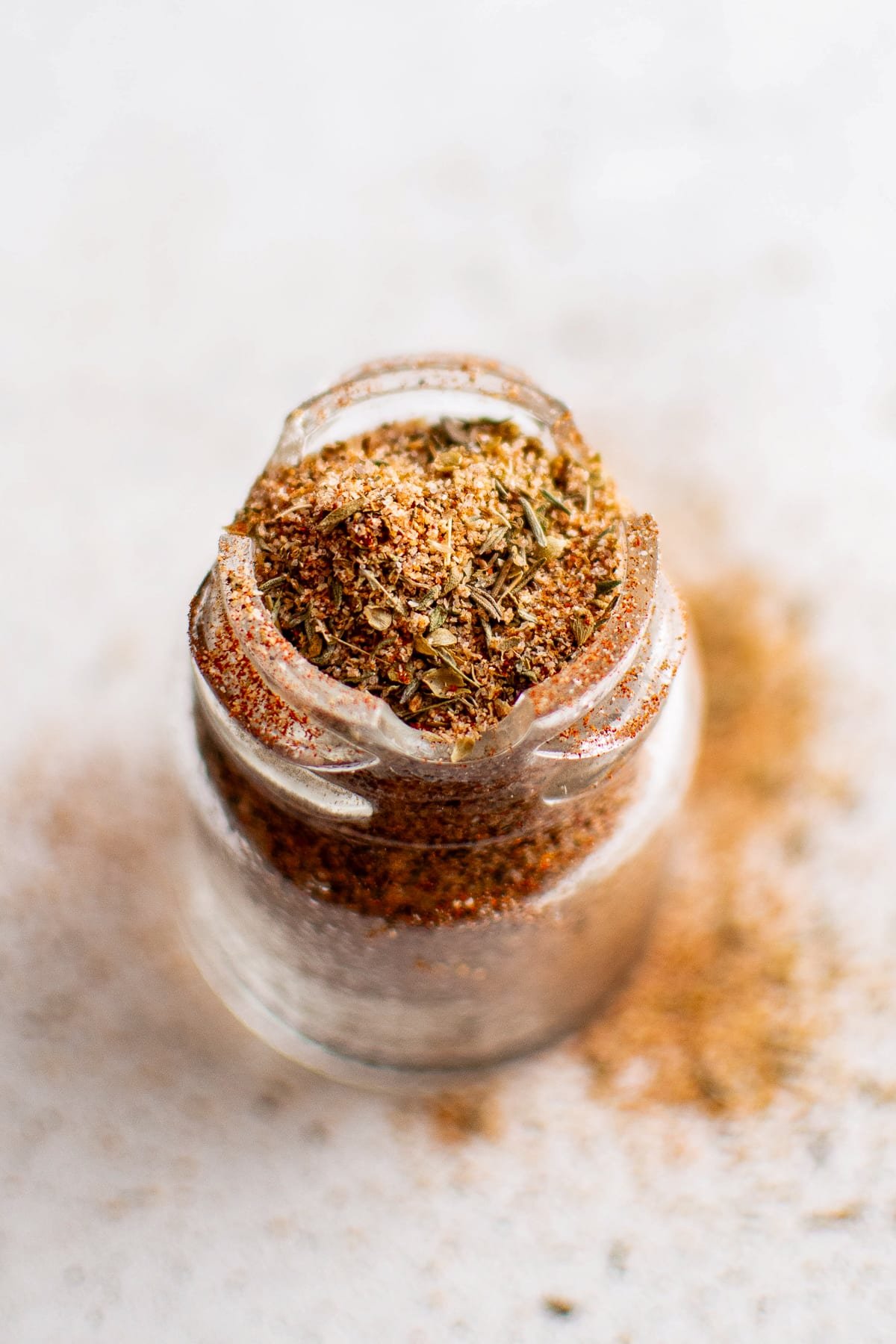 A small glass spice jar filled with homemade blackened seasoning. The mix is slightly spilling over the rim, and the jar is surrounded by scattered spices, emphasizing freshness and texture.