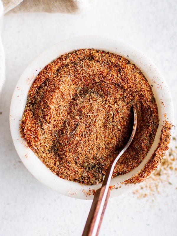 Overhead view of a white bowl filled with Creole seasoning and a spoon resting inside, surrounded by scattered spice.