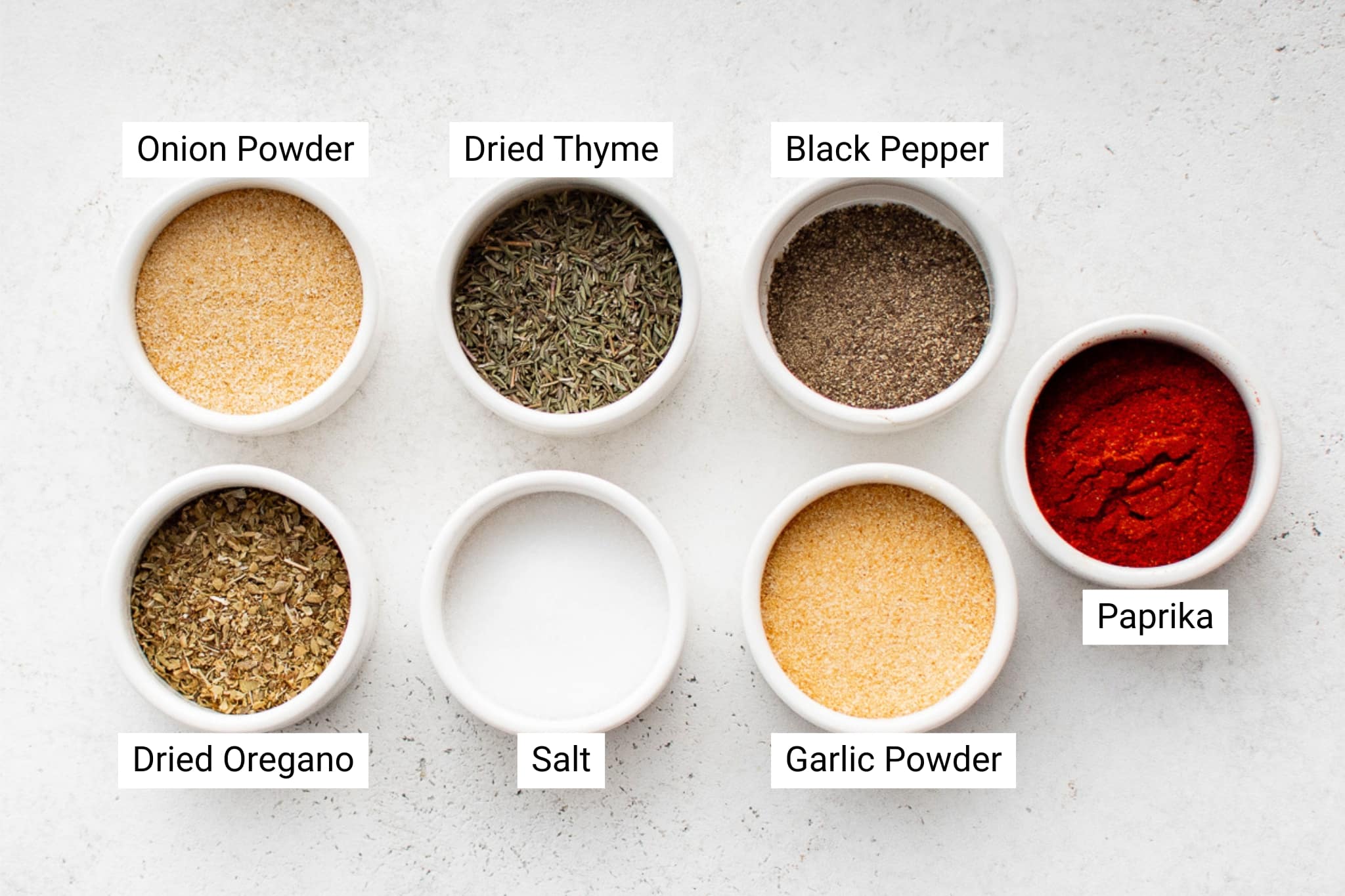 Overhead image of eight white bowls filled with labeled spices used to make blackened seasoning. Each bowl contains a single spice: onion powder, dried thyme, black pepper, paprika, garlic powder, salt, and dried oregano.