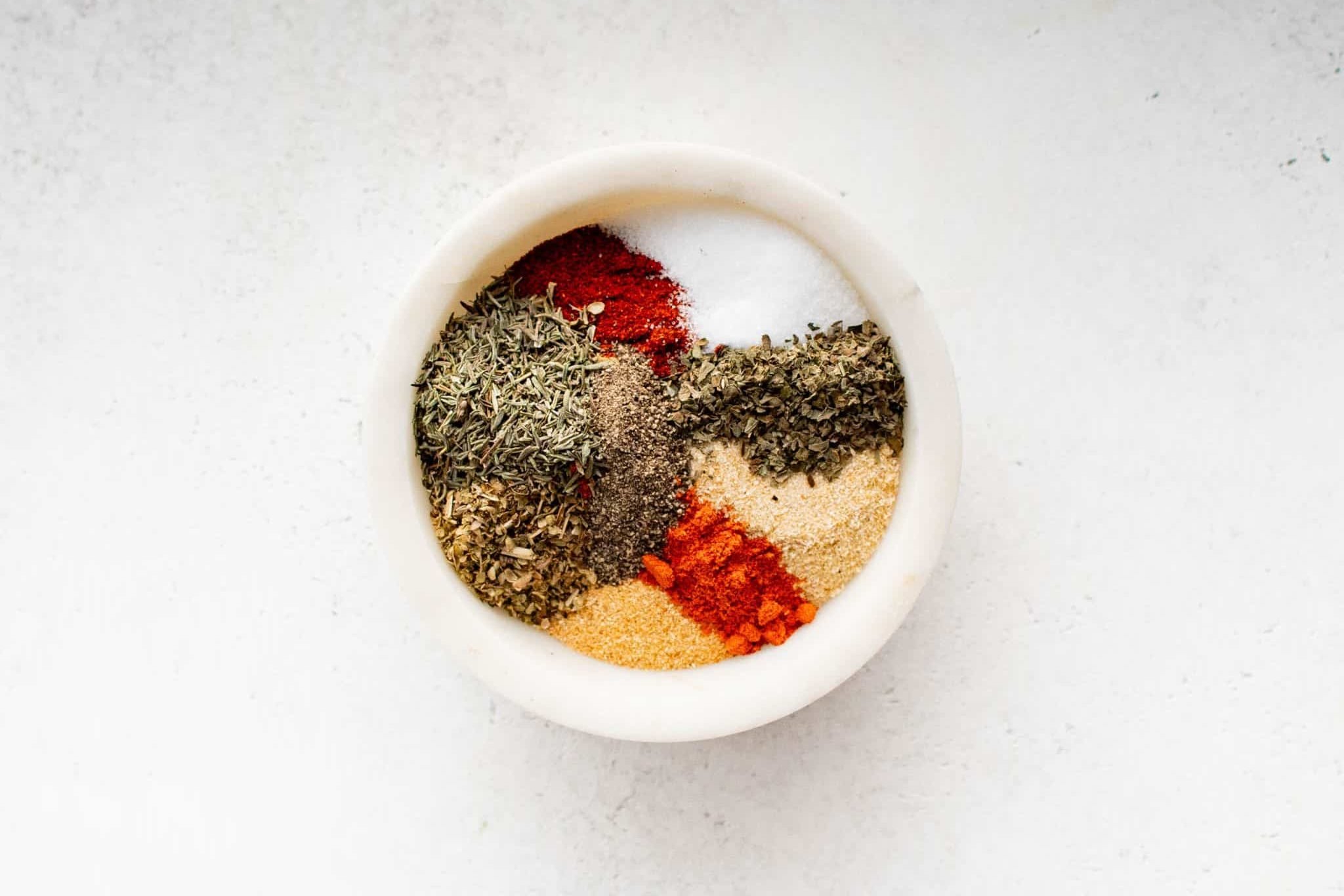 Top-down view of a white bowl filled with separated piles of herbs and spices for Creole seasoning, not yet mixed together.