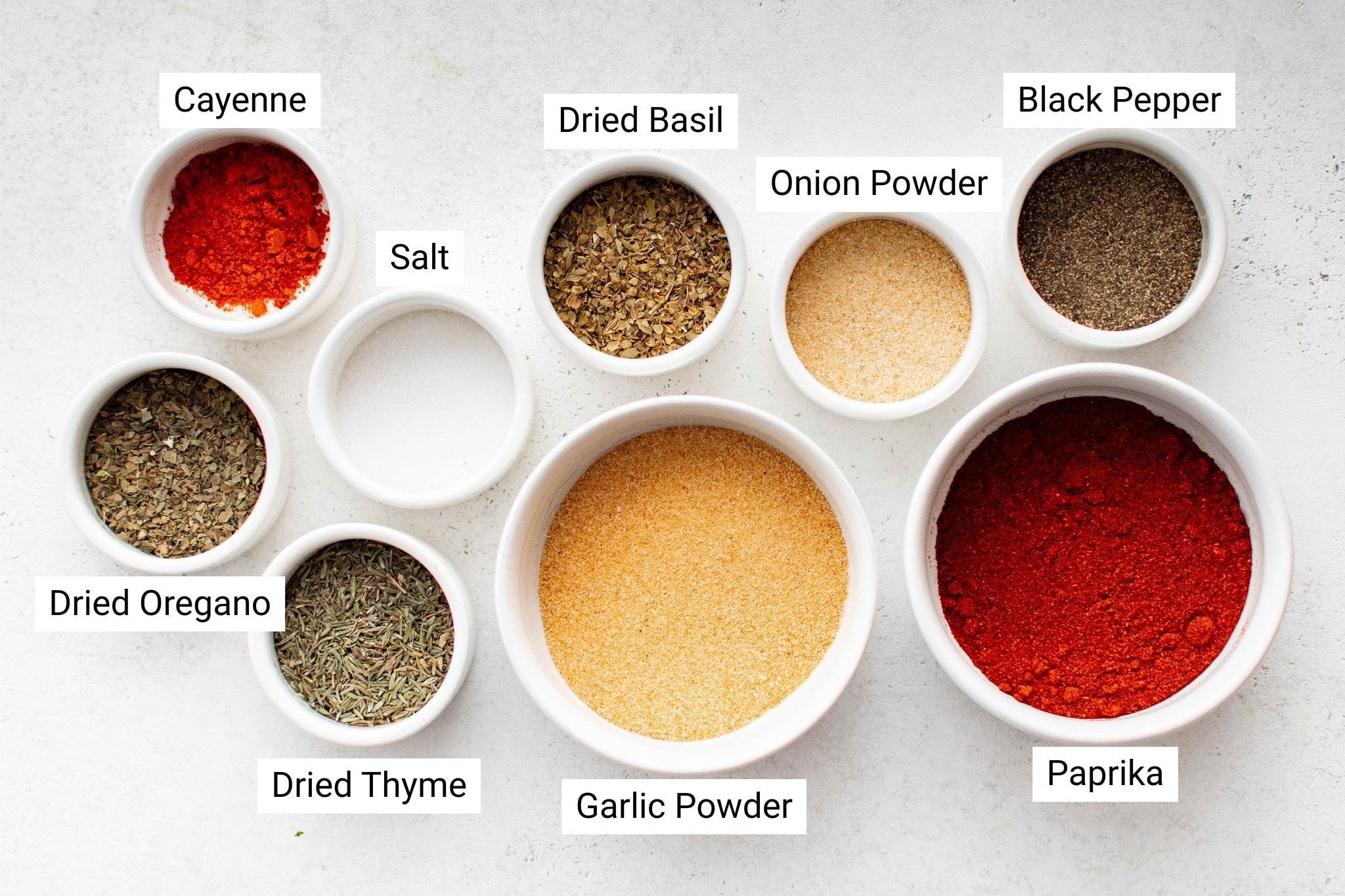 Overhead view of small white bowls filled with labeled spices and herbs used to make homemade Creole seasoning, including paprika, cayenne, garlic powder, onion powder, dried oregano, thyme, basil, salt, and black pepper.