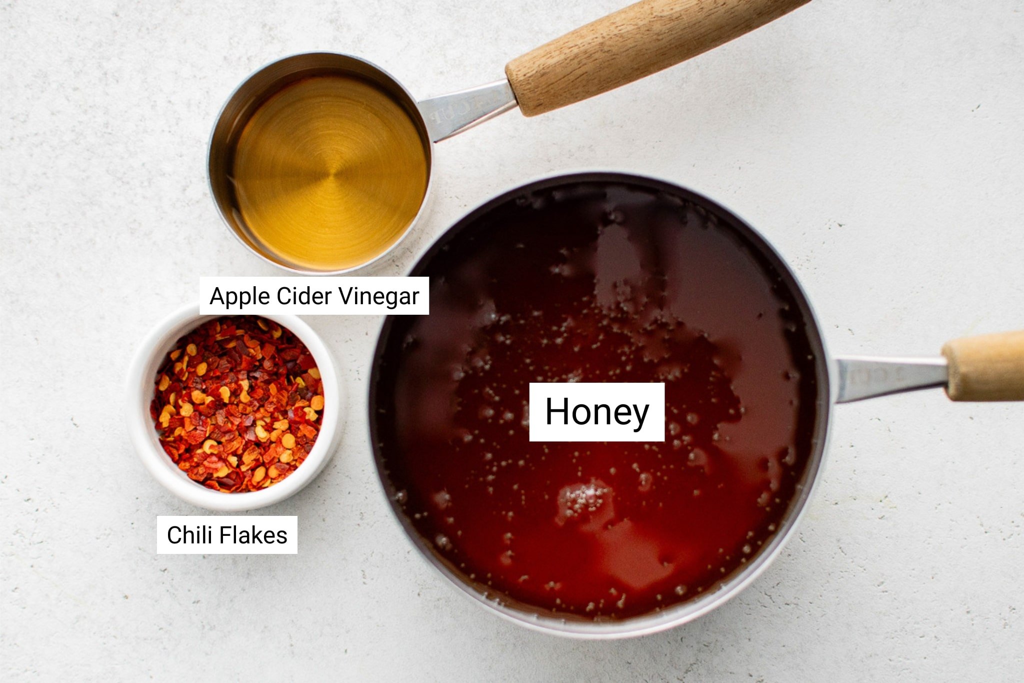 Overhead image showing the three labeled ingredients for hot honey: a white ramekin of crushed chili flakes, a metal measuring cup filled with apple cider vinegar, and a saucepan filled with honey.