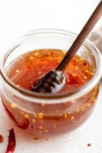 Close-up of a clear glass jar filled with golden hot honey, chili flakes suspended throughout, and a wooden honey dipper resting inside.