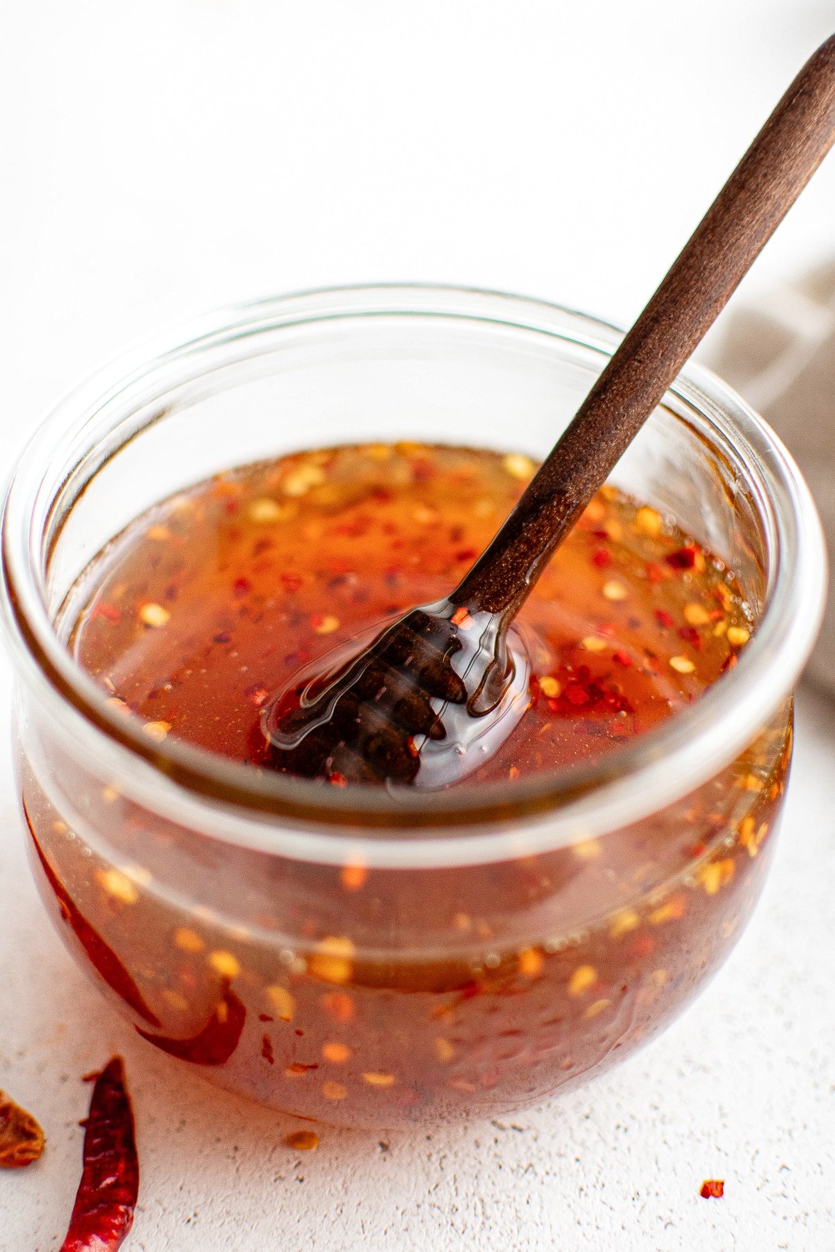 Close-up of a clear glass jar filled with golden hot honey, chili flakes suspended throughout, and a wooden honey dipper resting inside.