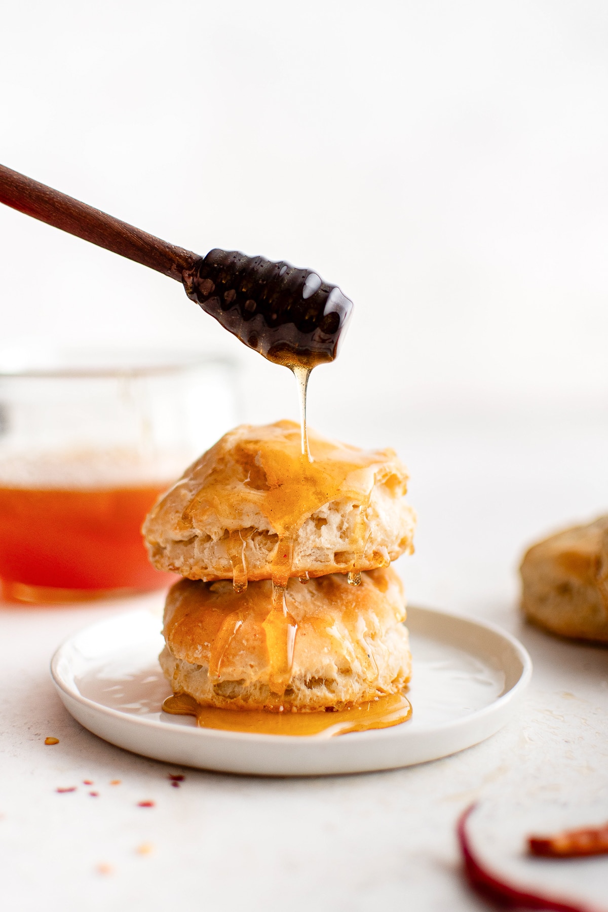 A close-up of strained hot honey being drizzled over stacked biscuits using a dark wooden honey dipper, with the jar of honey blurred in the background.
