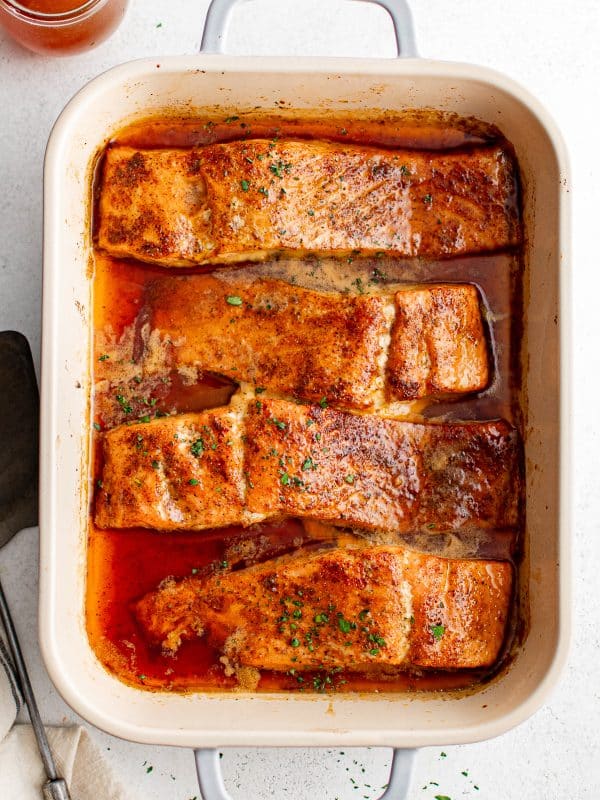 Baked salmon fillets garnished with fresh herbs and hot honey glaze in a cream-colored baking dish, viewed from above.