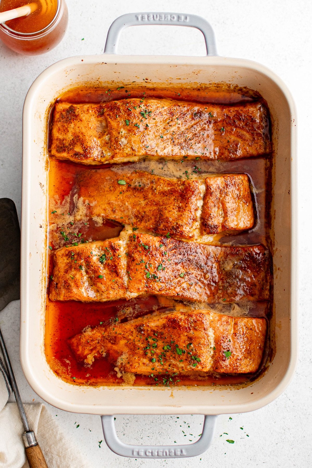 Baked salmon fillets garnished with fresh herbs and hot honey glaze in a cream-colored baking dish, viewed from above.