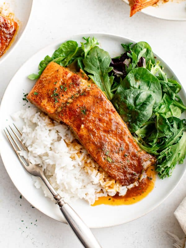 A baked salmon fillet served on a plate with white rice and mixed greens, topped with hot honey glaze.