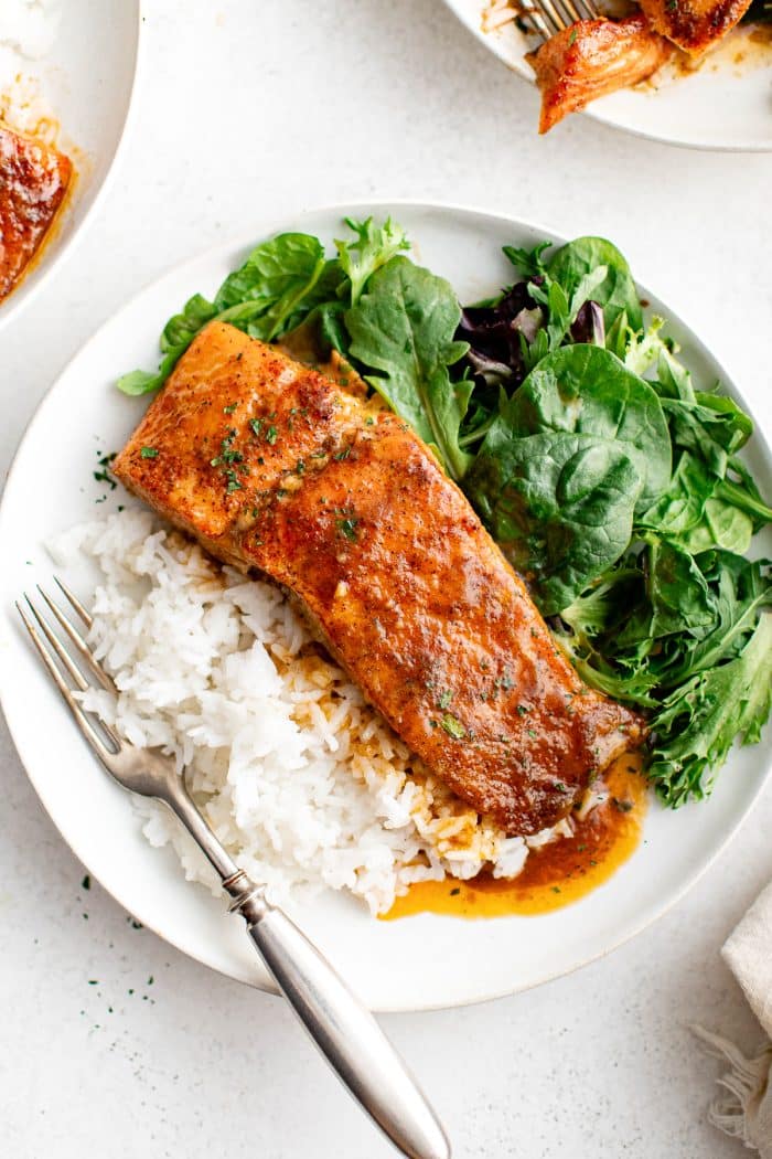 A baked salmon fillet served on a plate with white rice and mixed greens, topped with hot honey glaze.