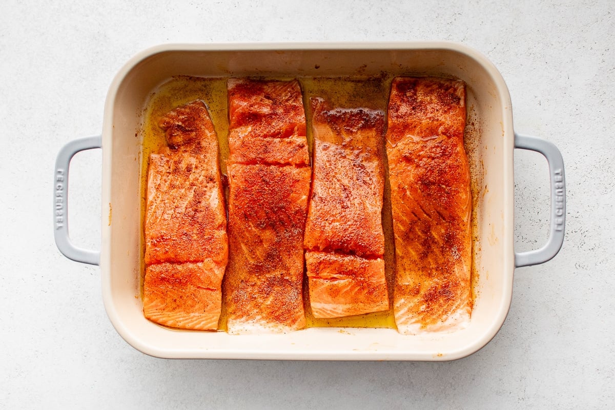 Four raw salmon fillets coated in a reddish-brown seasoning blend, placed in a cream-colored baking dish with olive oil.
