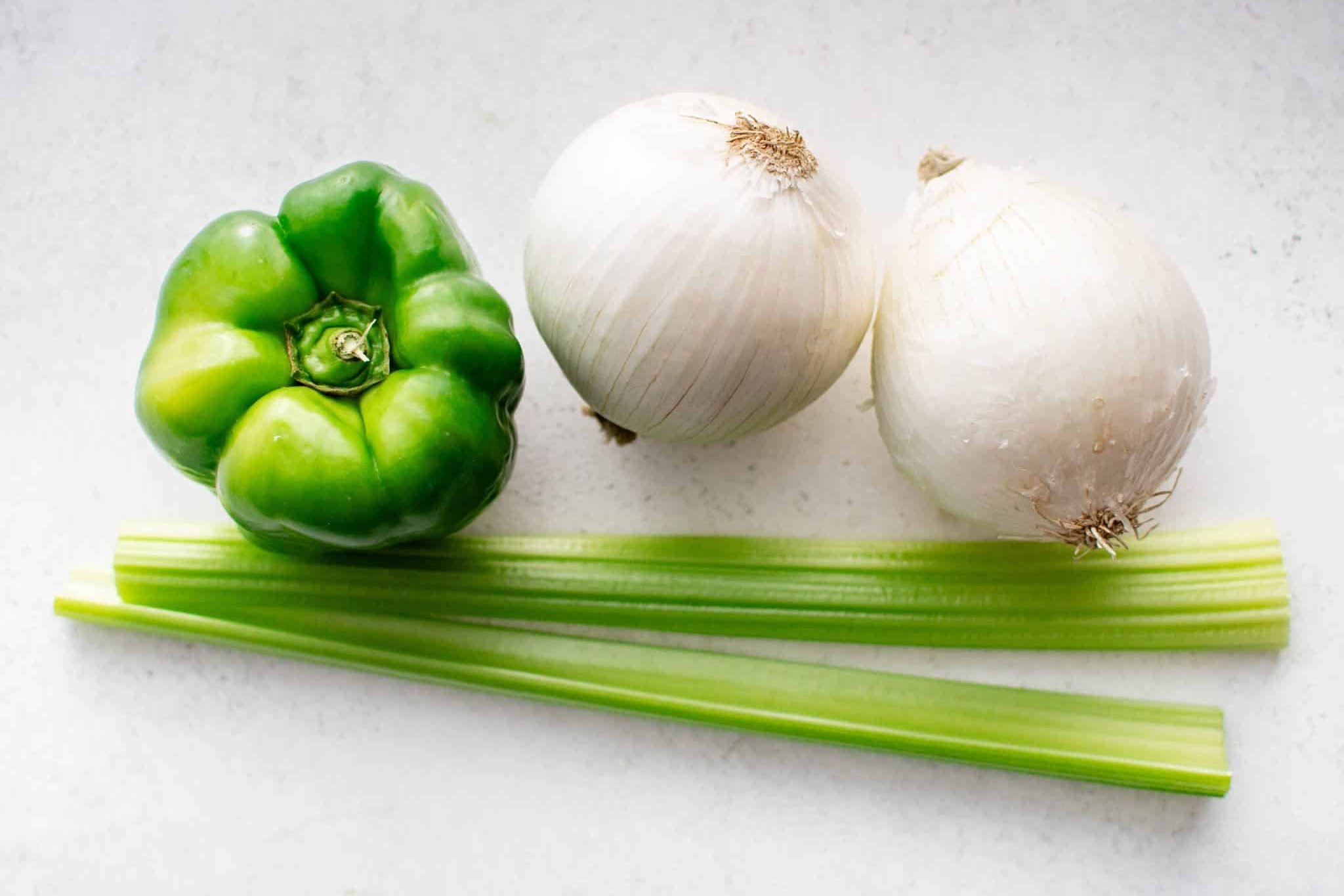 One green bell pepper, two celery stalks, and two white onions on a white surface.