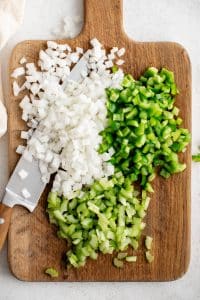 Diced onion, green bell pepper, and celery on a wooden cutting board with a knife.