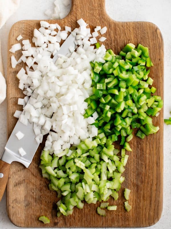Diced onion, green bell pepper, and celery on a wooden cutting board with a knife.