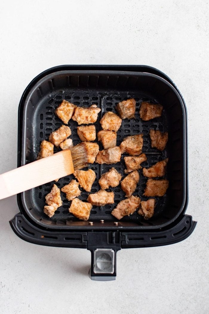 Partially cooked salmon cubes being brushed with teriyaki glaze inside an air fryer basket.