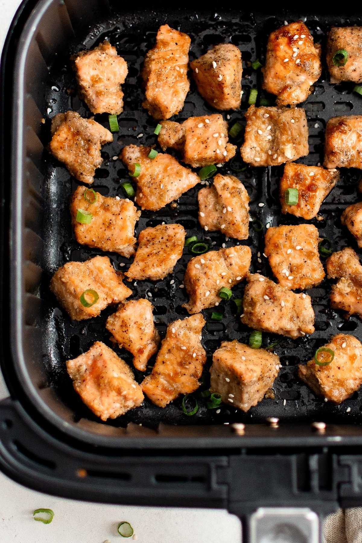Cooked salmon bites in an air fryer basket, garnished with sesame seeds and sliced green onions.