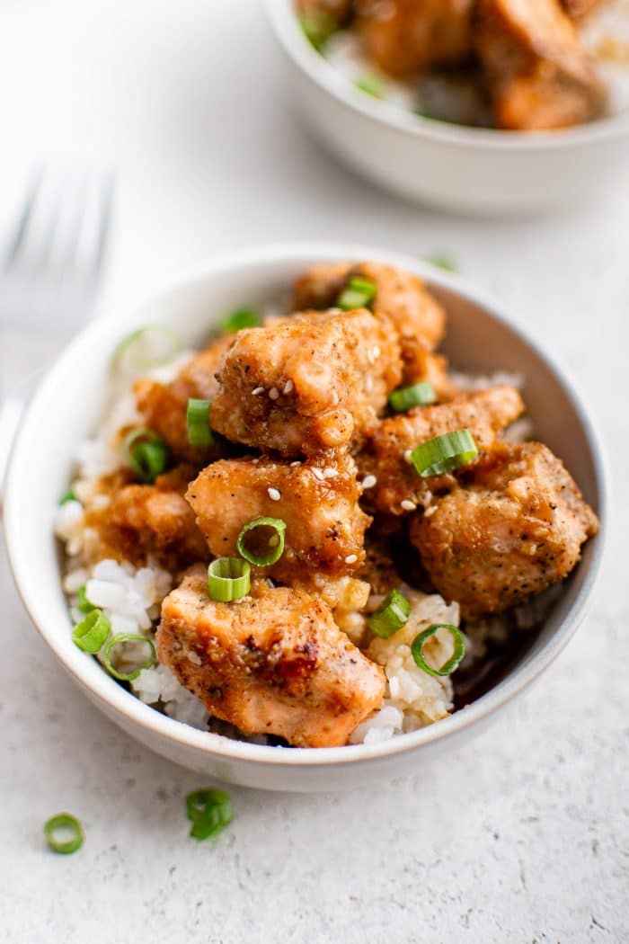 Bowl of white rice topped with golden-brown salmon bites, garnished with sesame seeds and sliced green onions.