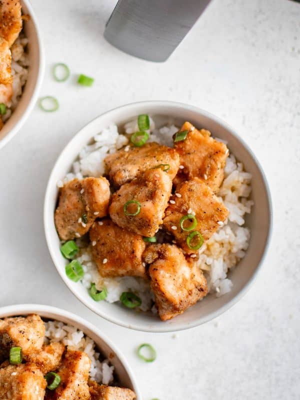 Overhead view of bowls filled with white rice and golden salmon bites, garnished with sesame seeds and sliced green onions.