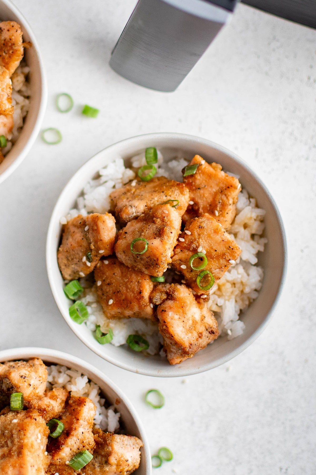 Overhead view of bowls filled with white rice and golden salmon bites, garnished with sesame seeds and sliced green onions.
