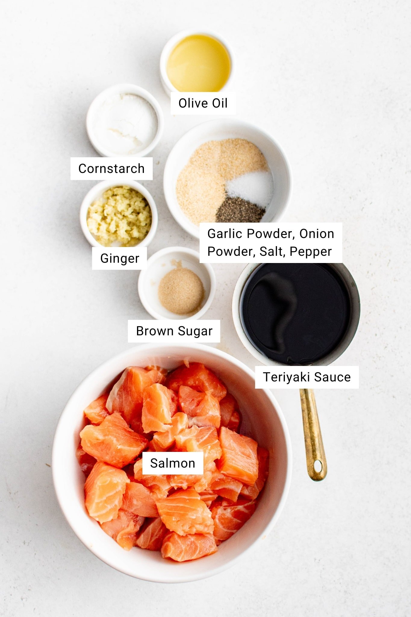 An overhead shot displays all the measured ingredients in small bowls on a white surface. Labels identify each ingredient, including a bowl of cubed salmon, a measuring cup of teriyaki sauce, and smaller bowls containing olive oil, cornstarch, minced ginger, brown sugar, and seasoning blends.