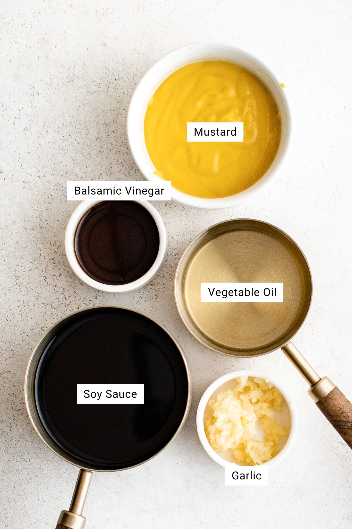 Overhead view of small bowls filled with steak marinade ingredients: soy sauce, balsamic vinegar, mustard, vegetable oil, and minced garlic, each labeled with text.