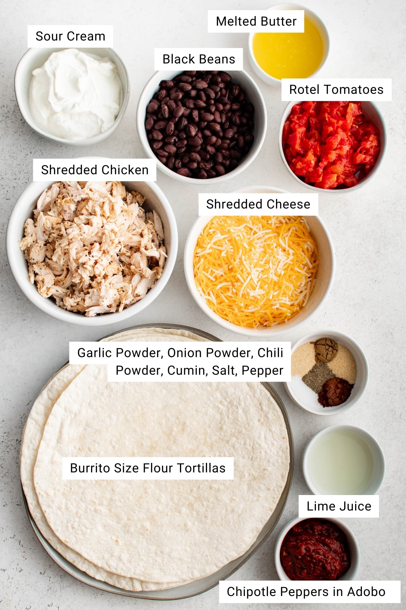 Overhead view of labeled ingredients for sheet pan quesadillas arranged neatly in bowls and plates. The items include sour cream in a small white bowl, melted butter in a small dish, black beans in a medium bowl, diced Rotel tomatoes in another bowl, shredded chicken in a larger bowl, shredded Mexican blend cheese in a bowl, a plate stacked with large burrito-size flour tortillas, a small dish of assorted spices including garlic powder, onion powder, chili powder, cumin, salt, and pepper, lime juice in a small bowl, and chipotle peppers in adobo in a small dish. Each ingredient is clearly labeled with text.