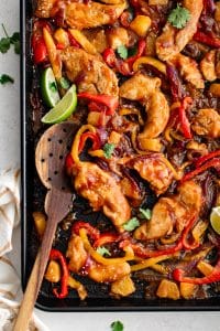 Juicy chicken tenderloins, roasted bell peppers, red onions, and pineapple chunks coated in Hawaiian sauce on a black sheet pan.