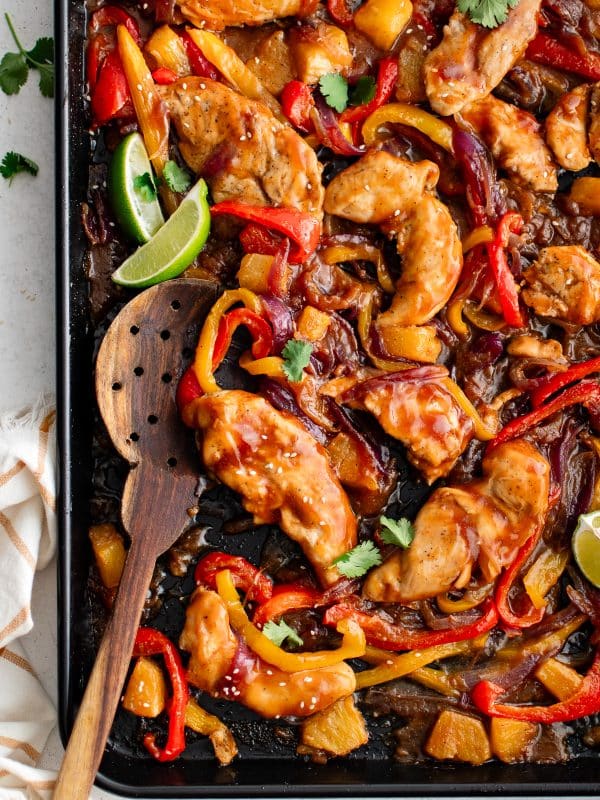 Juicy chicken tenderloins, roasted bell peppers, red onions, and pineapple chunks coated in Hawaiian sauce on a black sheet pan.