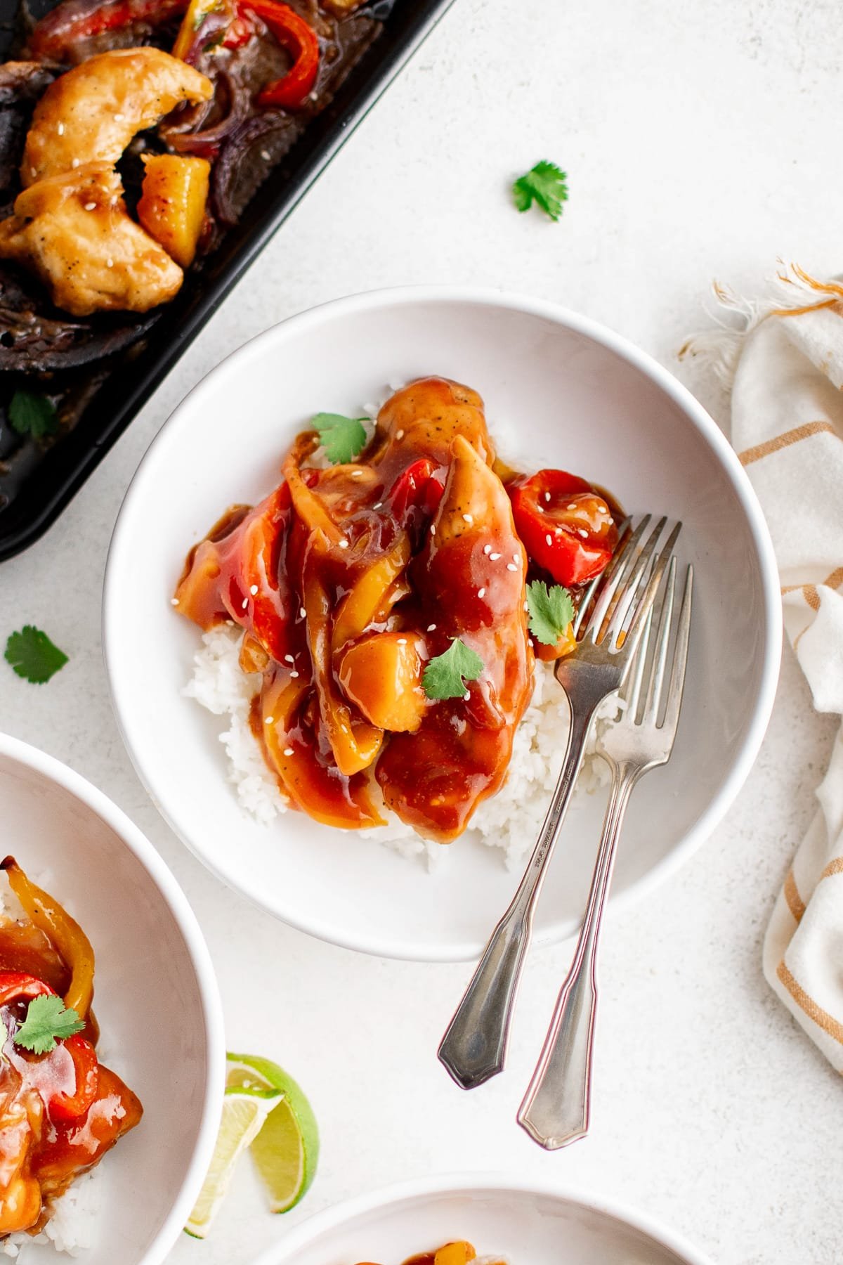 Bowl of white rice topped with saucy Hawaiian Chicken, pineapple chunks, and bell peppers, garnished with cilantro and sesame seeds, with a fork and spoon resting on the side.