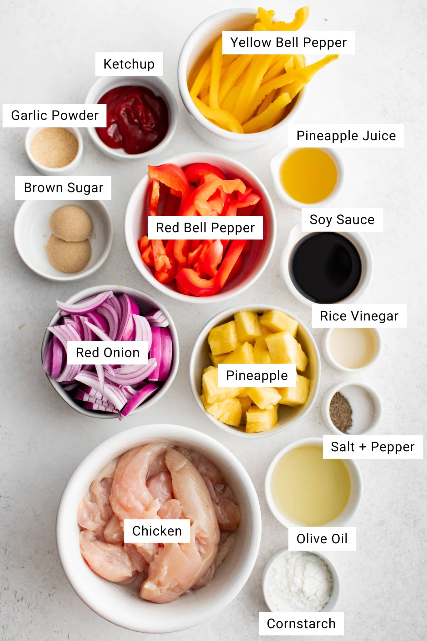 Overhead view of labeled ingredients for Hawaiian Chicken, including raw chicken tenderloins, pineapple chunks, red and yellow bell pepper strips, red onion slices, soy sauce, pineapple juice, rice vinegar, olive oil, cornstarch, brown sugar, ketchup, garlic powder, salt, and pepper.