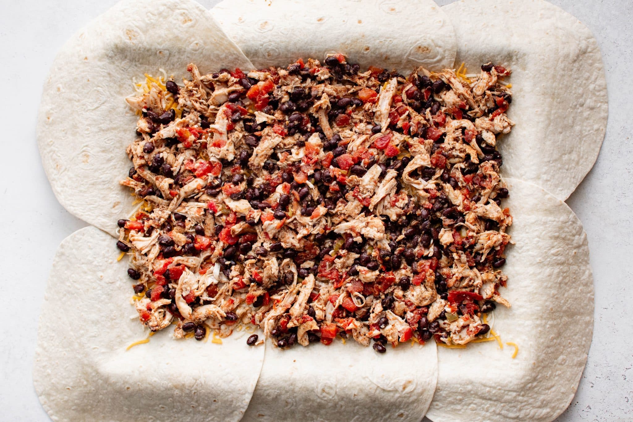 Overhead view of the chicken, black bean, and tomato mixture spread evenly across the shredded cheese on top of the tortillas.
