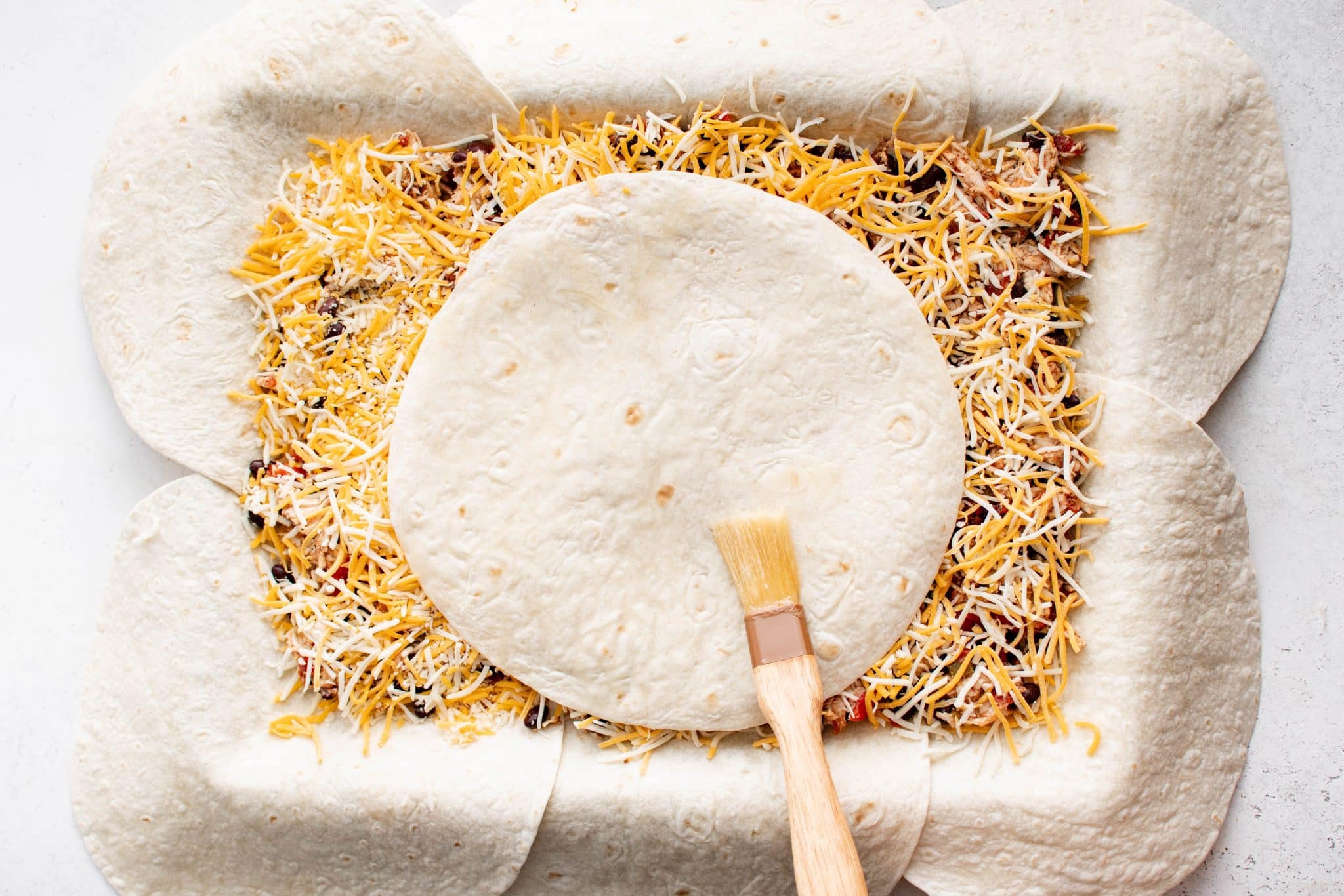Overhead view of an additional tortilla placed on top of the filling and cheese in the center of the pan, with a pastry brush applying melted butter.