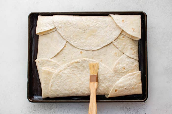 Overhead view of a sheet pan filled with folded tortillas covering the quesadilla filling, being brushed with melted butter using a pastry brush.