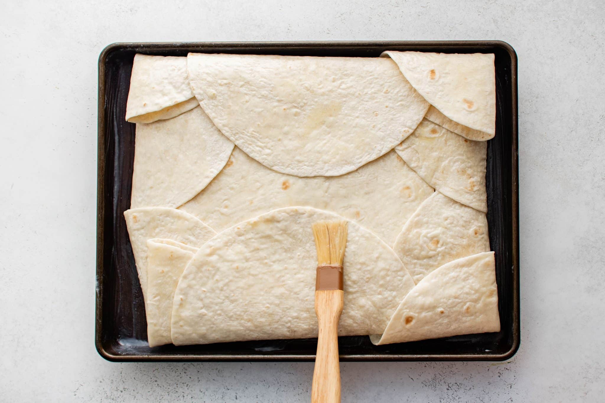 Overhead view of a sheet pan filled with folded tortillas covering the quesadilla filling, being brushed with melted butter using a pastry brush.