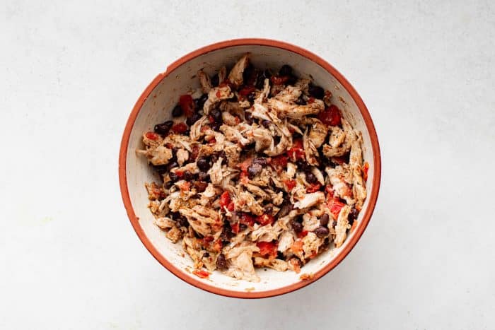 Overhead view of the same mixing bowl after the shredded chicken, black beans, tomatoes, and spices have been combined into a seasoned filling for sheet pan quesadillas.