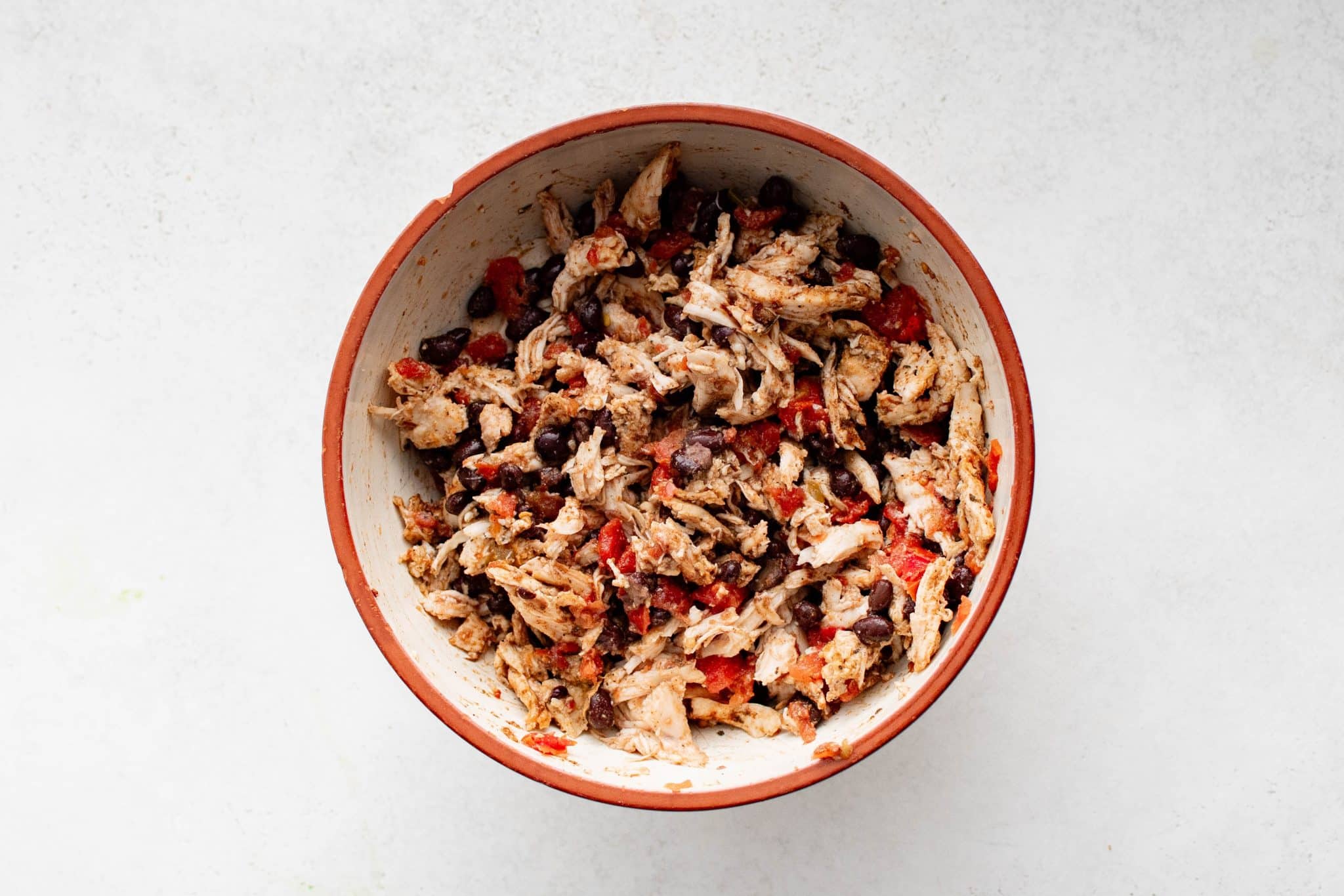 Overhead view of the same mixing bowl after the shredded chicken, black beans, tomatoes, and spices have been combined into a seasoned filling for sheet pan quesadillas.