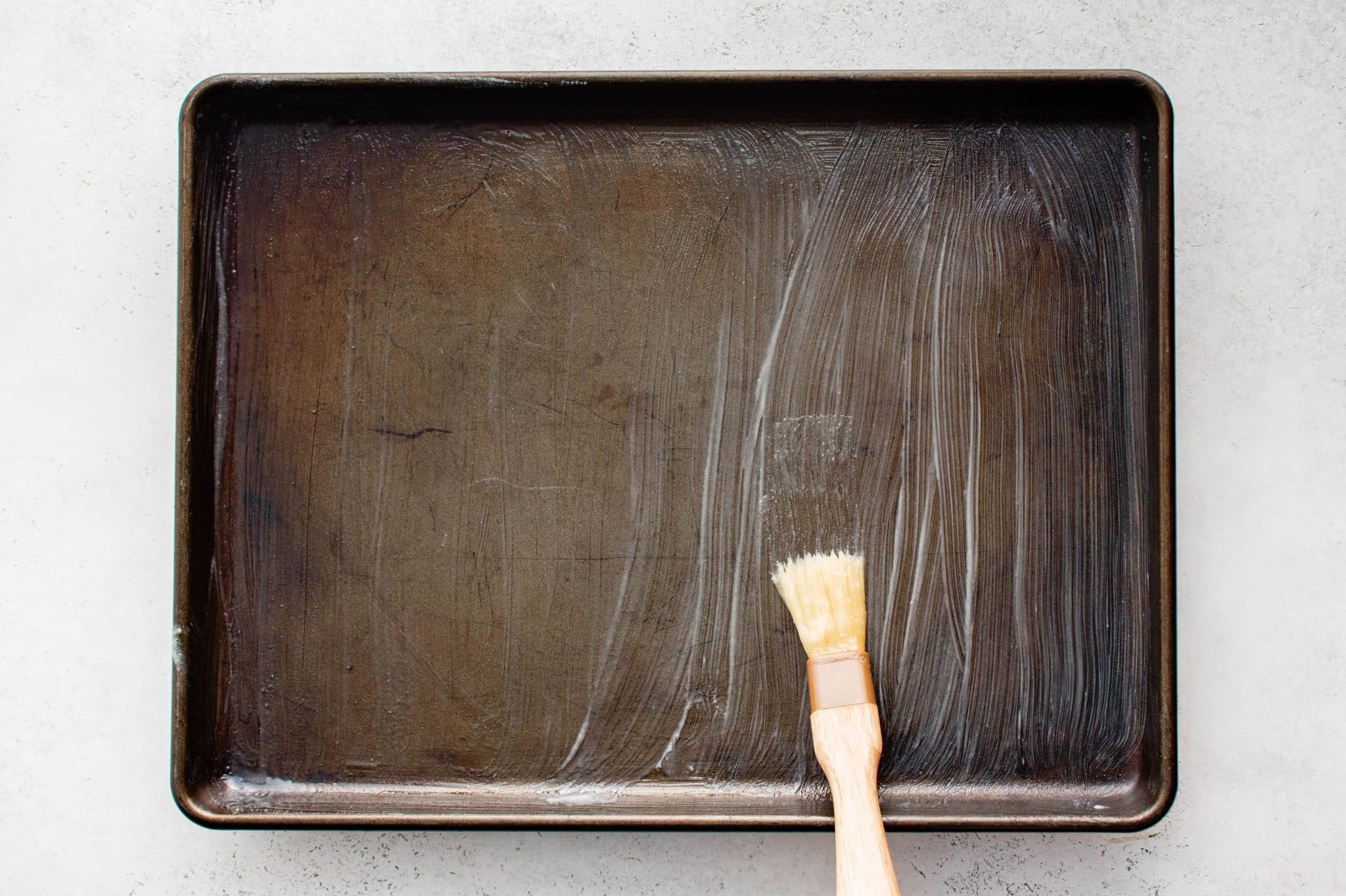 Close-up of a large metal baking sheet being brushed with melted butter using a pastry brush to prepare the pan for tortillas.