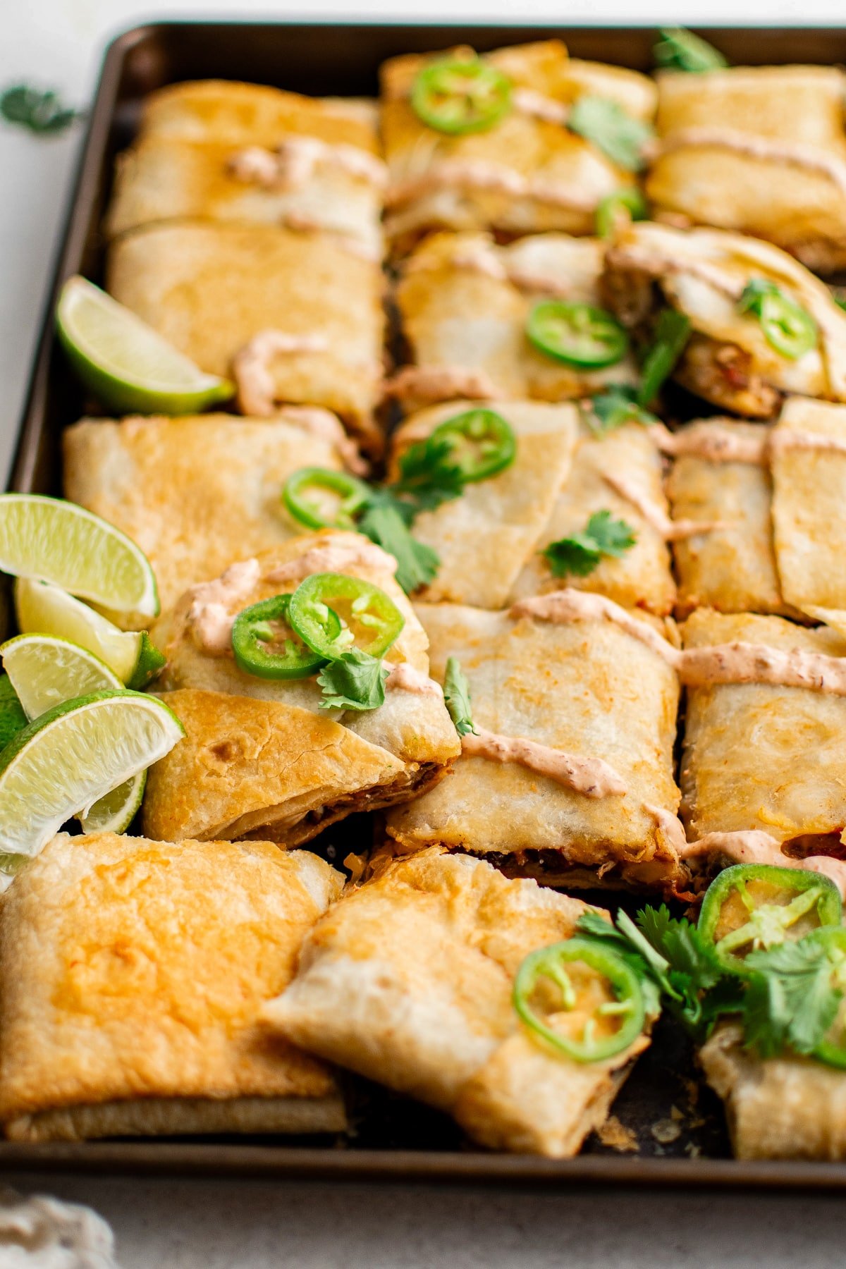 Close-up of golden-brown sheet pan quesadillas cut into squares, topped with chipotle crema, fresh jalapeño slices, and cilantro, with lime wedges arranged on the side for serving.