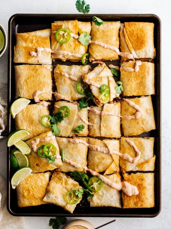 An overhead view of a full sheet pan of golden-brown quesadillas, cut into squares, topped with chipotle crema, fresh jalapeño slices, and cilantro, served with lime wedges and a bowl of chipotle crema on the side.