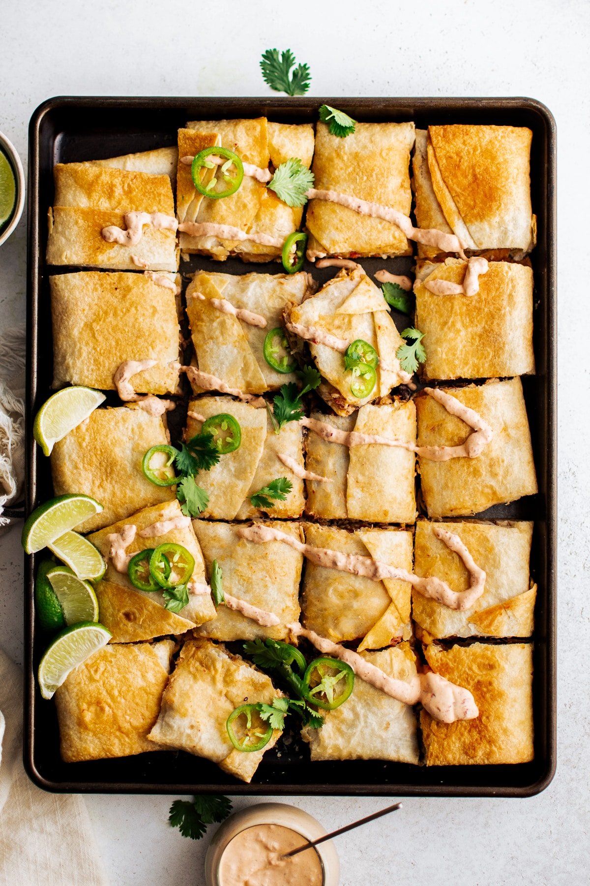 An overhead view of a full sheet pan of golden-brown quesadillas, cut into squares, topped with chipotle crema, fresh jalapeño slices, and cilantro, served with lime wedges and a bowl of chipotle crema on the side.