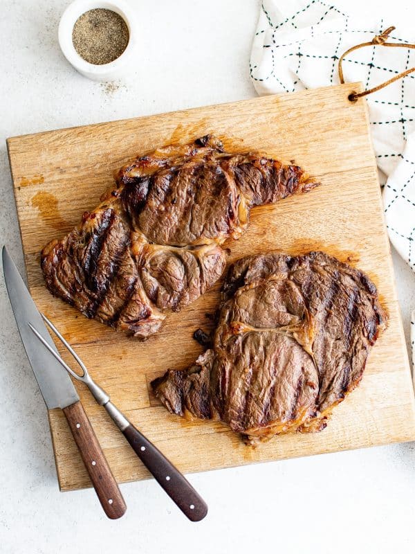 Two grilled steaks resting on a wooden cutting board with a carving fork and knife beside them, showing grill marks and juicy browned crust.