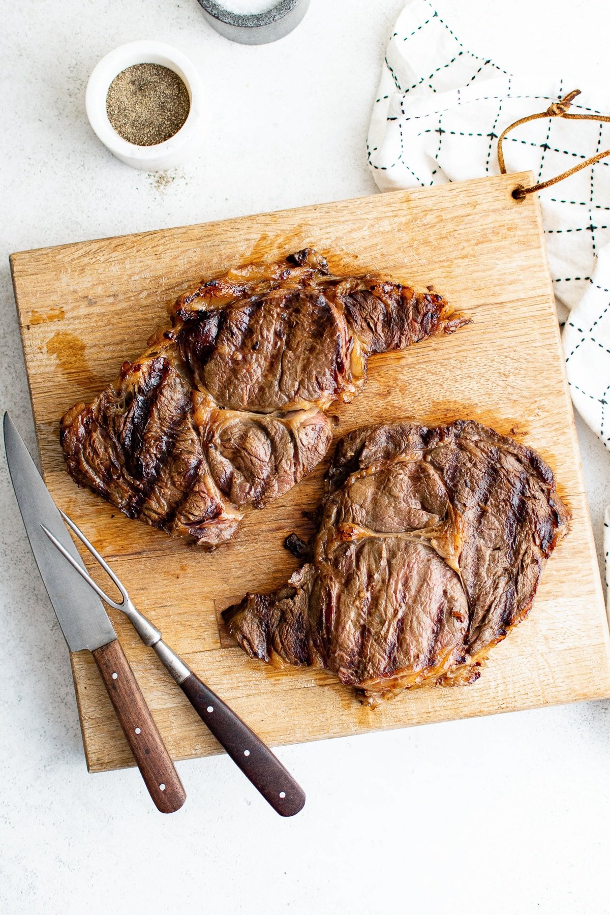Two grilled steaks resting on a wooden cutting board with a carving fork and knife beside them, showing grill marks and juicy browned crust.