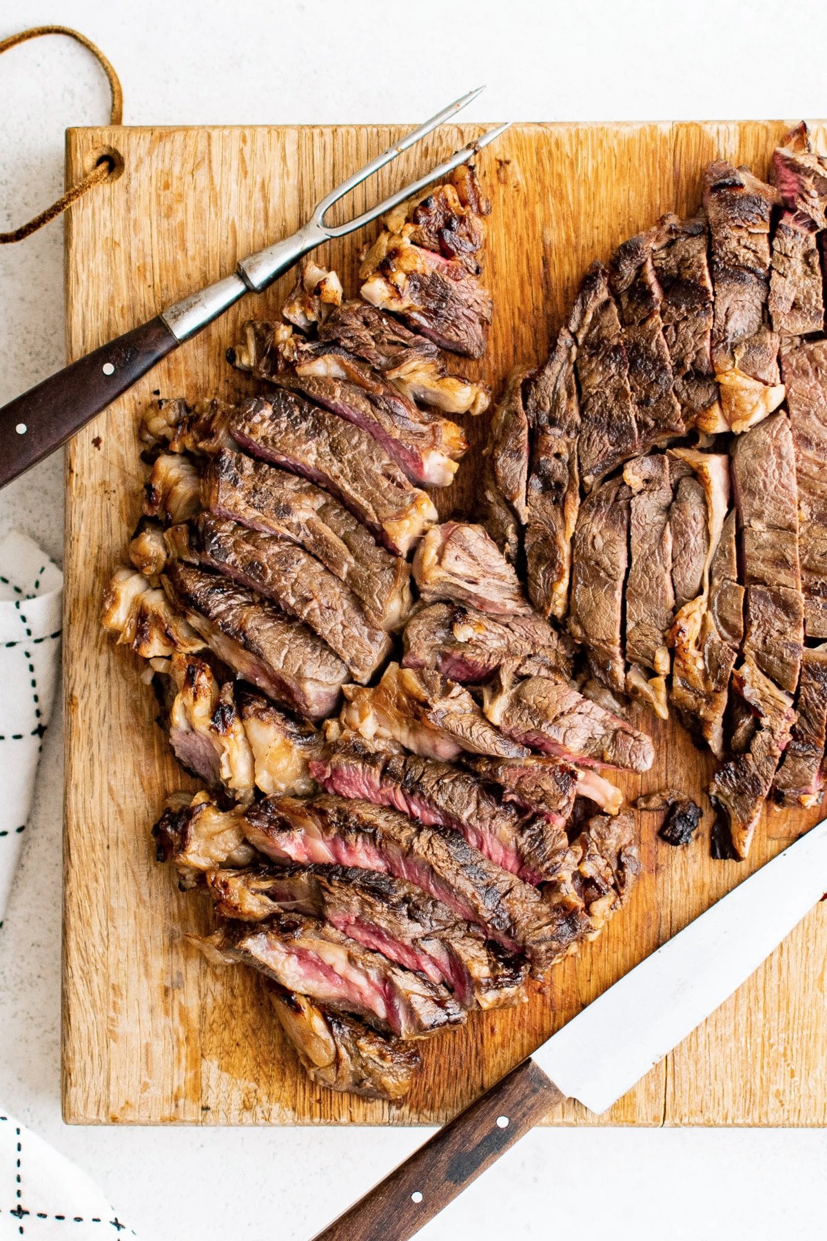 Sliced steak arranged on a wooden cutting board beside a carving knife and fork, showing pink center and juicy grilled exterior.