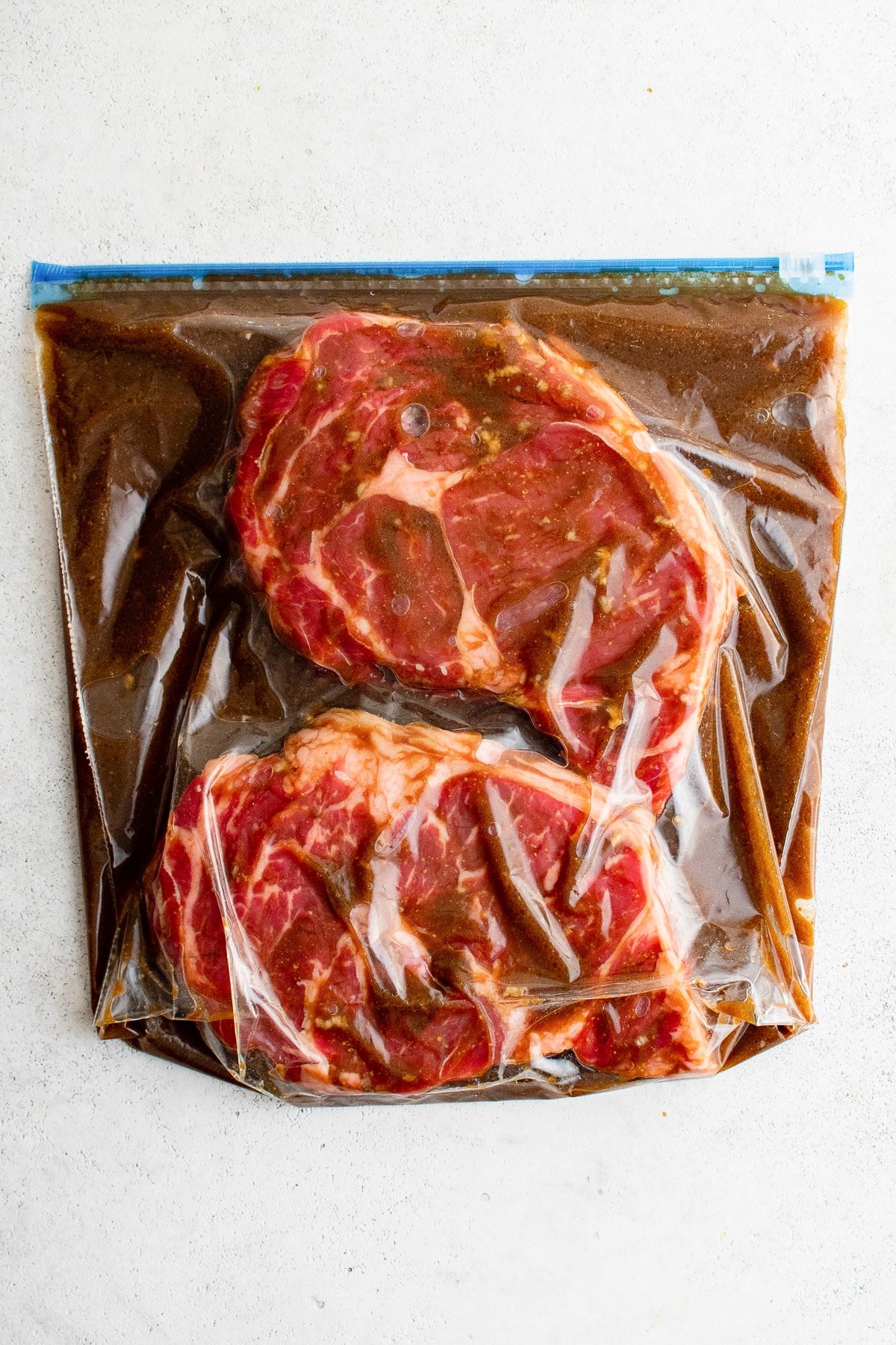 Two raw steaks fully coated in marinade inside a sealed plastic zip-top bag, laid flat on a white surface for even marinating. 