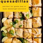 Overhead view of baked sheet pan quesadillas cut into squares, topped with sliced jalapeรฑos, cilantro, and chipotle crema, with lime wedges on the side. A yellow banner overlay reads, 'Easy Sheet Pan Quesadillas, perfect for game day or busy weeknight dinners.'