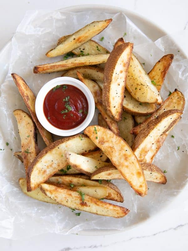Golden brown air-fried potato wedges served on a round plate lined with parchment paper, with a small bowl of ketchup for dipping.